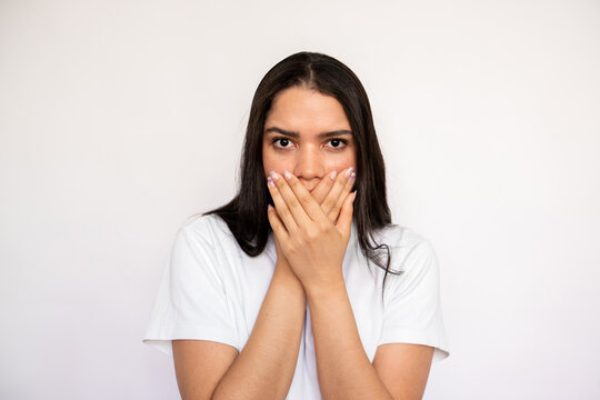 Portrait of scared young woman covering mouth with hand over white background. Caucasian lady wearing white T-shirt looking at camera in fear. Shock and stress concept
