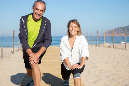 Close-up of happy aged couple exercising in summer. Smiling man and woman feeling great stretching legs on sandy beach looking at camera with joy. Active life and health care of aged people concept