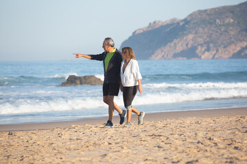 Man pointing finger into distance, walking on shore with wife. Long shot of cheerful senior couple...