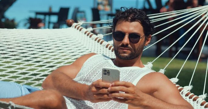 Side view of a handsome man with tanned skin on vacation, playing virtual online games on his mobile phone, while sunbathing in a hammock. Nice guy during summer vacation at coastal resort