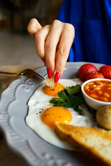 English breakfast, scrambled eggs with sausage, tomatoes, mushrooms, beans and toast. A plate of bright food, a juicy breakfast. Women's hands on the background