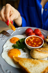 English breakfast, scrambled eggs with sausage, tomatoes, mushrooms, beans and toast. A plate of bright food, a juicy breakfast. Women's hands on the background