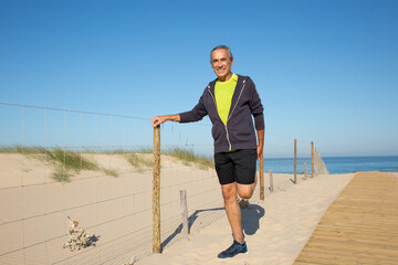 Portrait of smiling senior man preparing for training outdoors. Grey-haired man in casual clothes standing on sand near sea stretching legs with pleasure. Active lifestyle of elderly people concept
