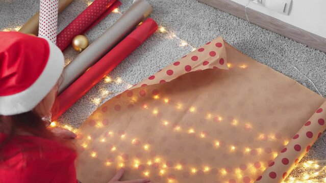 Girl in a Santa hat creates New Year's packages from red wrapping paper. Young woman is packing Christmas presents for her friends and relatives. Decorations, preparation for the winter holiday season
