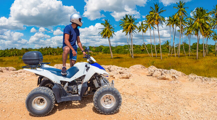 ATV driver in tropical area. Man with ATV examines palm trees. Concept of extreme tourism. Quad bike driver in helmet looks into distance. ATV racing in tropical area. Man with quad bike © Grispb