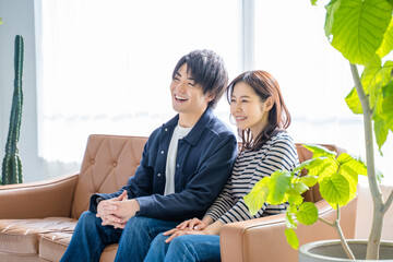 リビングのソファーに座りテレビ番組を鑑賞する笑顔の若い夫婦カップル