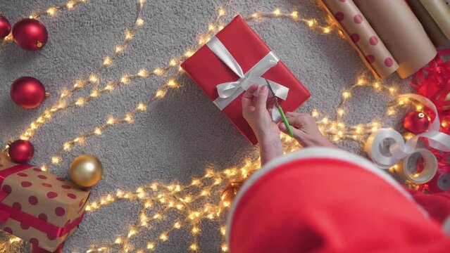Girl in a Santa hat creates New Year's packages from red wrapping paper. Young woman is packing Christmas presents for her friends and relatives. Decorations, preparation for the winter holiday season