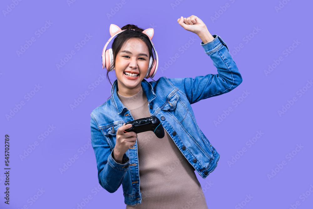 Wall mural Beautiful Asian young woman in jacket jean and playing video games using joysticks with headphones on voilet background isolated.