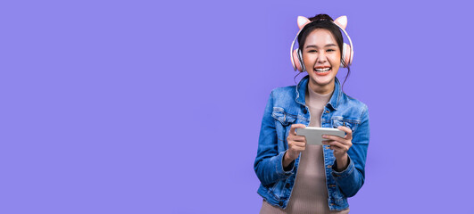Beautiful Asian young woman in jacket jean and playing video games using joysticks with headphones on voilet background isolated. - obrazy, fototapety, plakaty