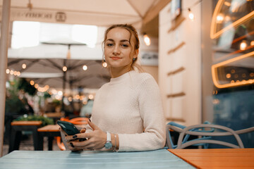 Young stylish woman in sweater using a smart phone standing outdoors on the street in Warsaw. Portrait of beautiful woman reading message on the street.