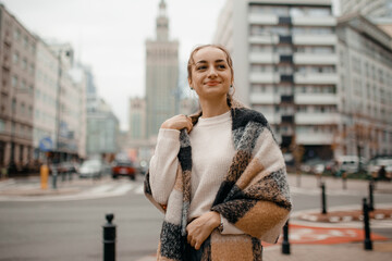 Happy young adult woman smiling with teeth smile outdoors and walking on city street weating winter clothes and knitted scarf. Female young traveler in Warsaw, Poland. - obrazy, fototapety, plakaty