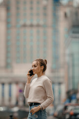 Fototapeta premium Young woman using mobile phone walking on the city street in Warsaw, Poland. Successful woman calling with cell telephone outside,business talking via mobile phone and walking on the city street.