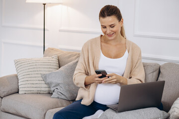 Pregnant woman sitting on coach using laptop and holding headphons by the belly