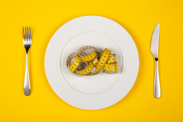 Flat lay image of tape measure resembling spaghetti on white plate with fork and knife near isolated on yellow background.