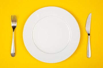 flat lay image of empty white plate, fork and knife on yellow background