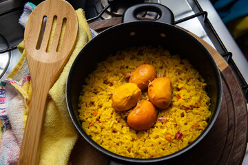 Uma panela preta com arroz com pequi, um pano de prato e uma colher de madeira, sobre uma tábua de madeira. Um prato típico brasileiro.