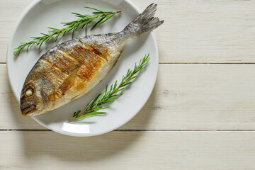 Dorado, cooked, grilled fish fried, two branches rosemary, on plate, on white wooden board background, top view, space to copy text.