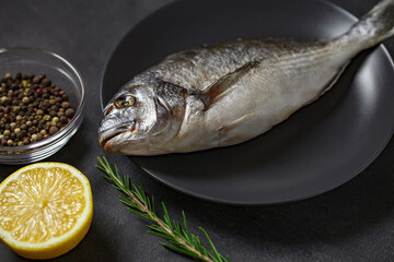 Dorado, raw fresh uncooked fish, lemon, rosemary, pepper mix, on dark background, side view, selective focus.