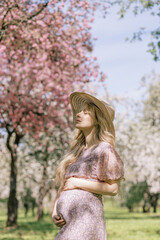 Pregnant young adult woman in a pink dress and a white cardigan in a spring blooming cherry garden. Sakura tree. Waiting for a baby