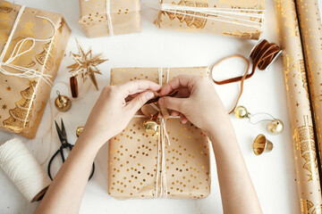 Hands wrapping stylish golden christmas gifts flat lay. Person preparing modern christmas gift boxes, festive paper, golden ornaments and thread composition on white wood. Merry Christmas!