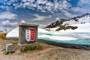 Obraz premium Monument of Flag and coat of arms of the canton of Valais with snowy Col du Nufenen mountain