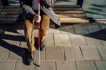 Close-up of a blind man with a walking stick sitting on a bench at a public transport stop