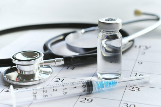 A medical syringe needle with plain unmarked vial of medicine laying on a calendar page with a stethoscope. Concept for scheduling childhood immunizations or covid-19 tetanus or flu shot boosters. 