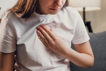 Young unhealthy female sit on couch having difficulty breathing pain of heart, touches his chest with hand. Trouble breathing, chest pain. Heart attack, thoracic osteochondrosis, panic attack concept - obrazy, fototapety, plakaty