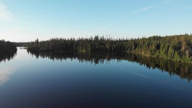 Drone moving sideways over a very calm boreal lake that is surrounded by forest. The still water is reflecting the clear blue sky
