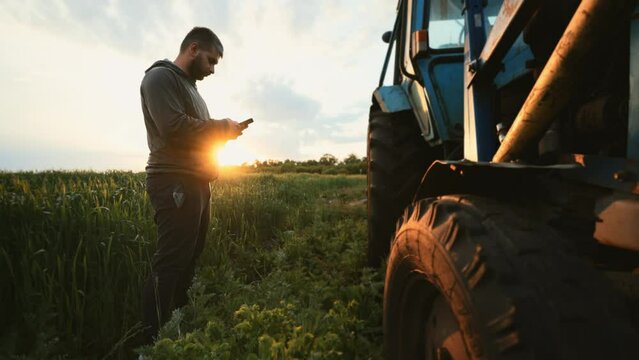 Farmer man stands in green field near tractor and uses mobile phone for text messaging or remote control of agriculture. Break rest in working day. Wireless technology. Communicate on social networks.