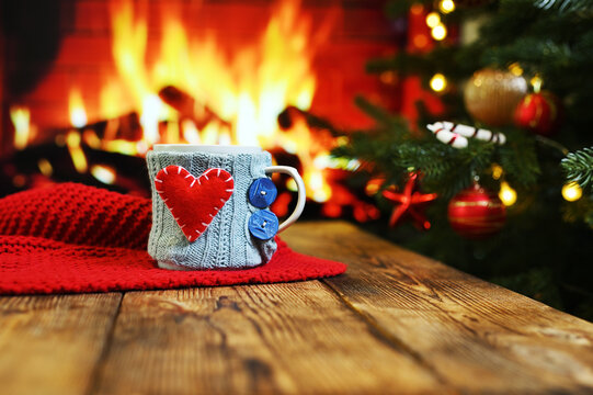 a mug in a knitted case with a red heart on the side on a wooden table against the background of a red plaid, a fireplace and a Christmas tree