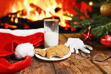 a bitten oatmeal cookie and an unfinished glass of milk on a wooden table against the background of Santa Claus clothes, a Christmas tree and a fireplace