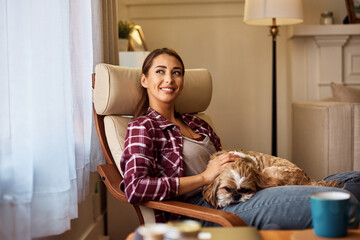 Happy woman looking through window while relaxing with her dog in armchair.