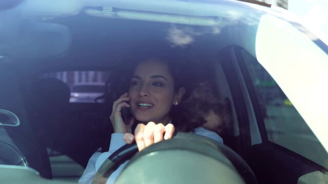Close up of happy young pretty woman driving a car and talking on smartphone in good mood. Caucasian attractive cheerful female speaking on mobile phone call while drives vehicle
