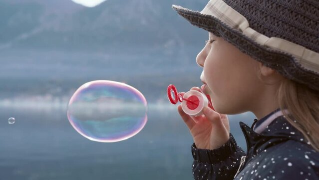 Kid blowing soap bubbles outdoors at winter sea beach. Child having fun on nature. Girl playing, enjoying holidays by lake. Concept of mindfulness, mental health, digital detox. Lifestyle moment