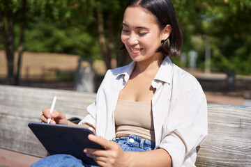 Portrait of young asian girl sitting in park with laptop, writing with digital pen or drawing, getting inspiration from outdoors