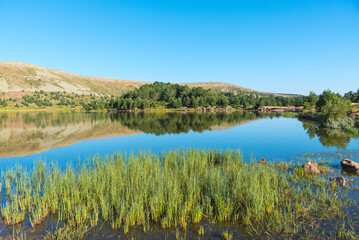 Landscape of a lake surrounded by covered greenery mountains reflected in the water under a blue sky, Neila Lagoons Natural Park, Burgos, Spain