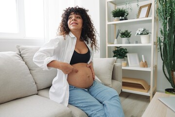 Pregnant woman smile and happiness sits on the couch freedom and strokes her belly feels kicks with the baby in the last month of pregnancy, mother's day lifestyle