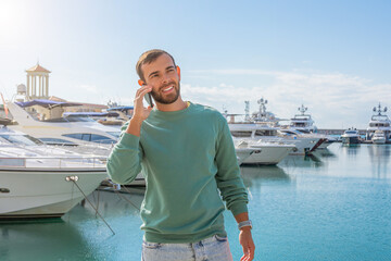 Caucasian male in green pullover is talking on phone in port against the backdrop of expensive yachts. Successful man business negotiations while traveling.