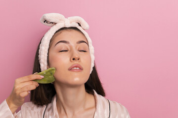 Face massage. beautiful young woman in pink pajamas does a facial massage with green quartz gouache...