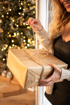 Beautiful young girl holding a Christmas gift wrapped in Kraft paper against a sparkling Christmas tree. Vertical image.