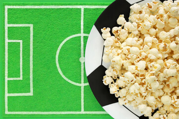 Popcorn on big plate painted like soccer ball and dish mat like football field made of green felt with place for text. Template of football soccer party table. Top view, copy space, mockup.