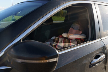 A tired driver sleeps in the car during the day, covered with a blanket and wearing a sleep mask over his eyes.