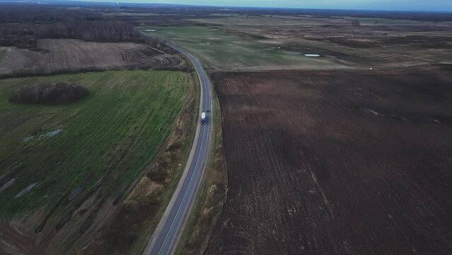 Car and truck drives along road near field, aerial view. Car in motion on asphalt road during trip, rural landscape. Automobile is driving on highway in autumn. Car moving in countryside along road.