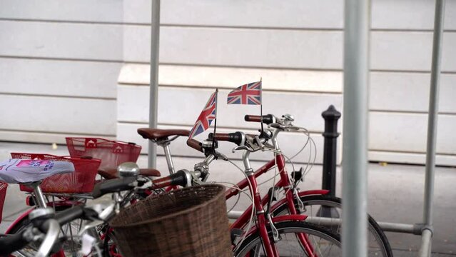 The British flag flutters in the wind on the handlebars of bicycles parked on the street of a European city. Red bikes are parked nearby.