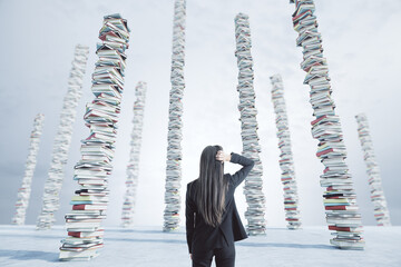Back view of young businesswoman looking at towers of books on sky background. Education, knowledge...