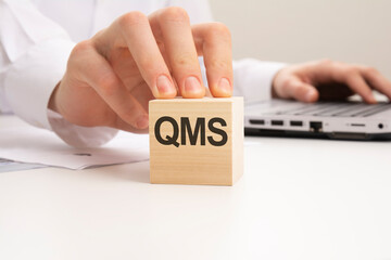 a wooden block with the letters QMS written on it on a white background. QMS - short for Quality...