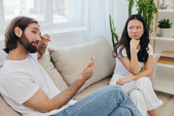 Fototapeta premium A man looks at the phone screen during an argument with his girlfriend. The angry and hurt woman looks in his direction and is sad. Family discord at home, phone addiction