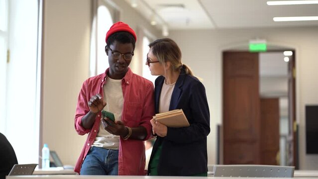 Two multiracial students, man and woman studying together, communicating in university library. African hipster guy holding smartphone explaining to Caucasian girl how to use app for borrowing books