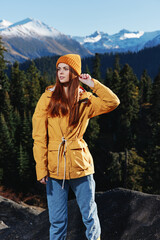 Woman in yellow raincoat with red hair on a hike stands in front of mountains in yellow cap autumn sunset light 